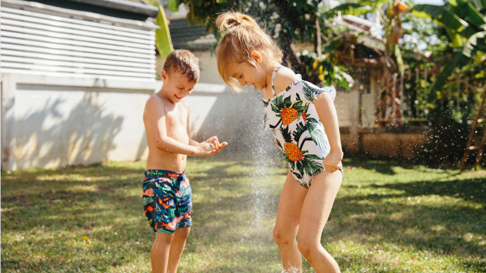 A little boy and girl wearing bright coloured swimwear are playing in a backyard sprinkler on a sunny day. They are smiling and running through the water. They applied SPF with their Sunscreen applicator by sunnybod.