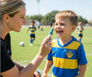 Mother reapplying sunscreen outdoors with a SUNNYBOD refillable sunscreen applicator to a smiling child wearing a soccer uniform on a sports field.