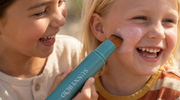 Two children laughing outdoors while one applies sunscreen to the other's face using a SUNNYBOD refillable sunscreen applicator brush