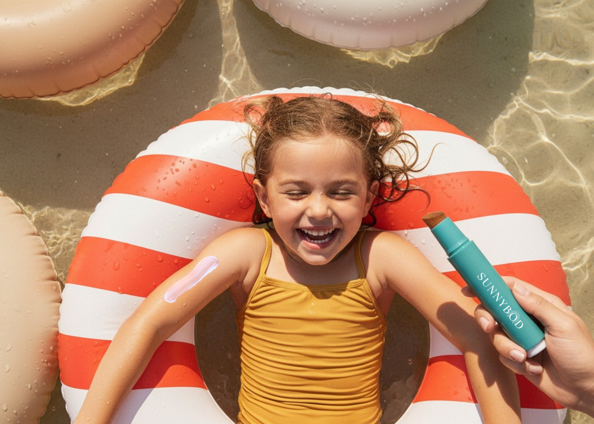 Child lying on a striped pool float in  the water, smiling broadly. At the right of frame an adult is holding a SUNNYBOD refillable sunscreen Applicator, used to apply SFP easily to their child. 
